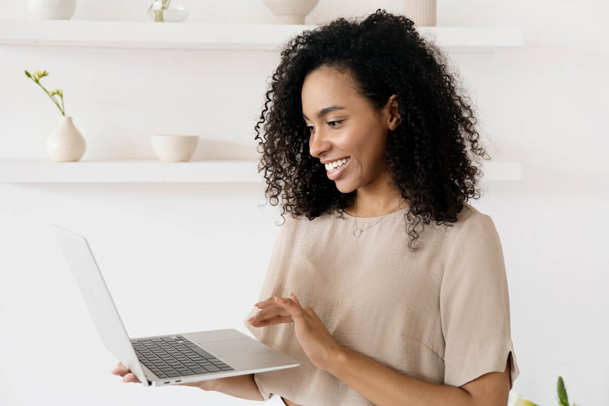 Girl standing up with shoulder-length curly dark hair, holding a laptop and looking down at it while smiling Girl standing up with shoulder-length curly dark hair, holding a laptop and looking down at it while smiling