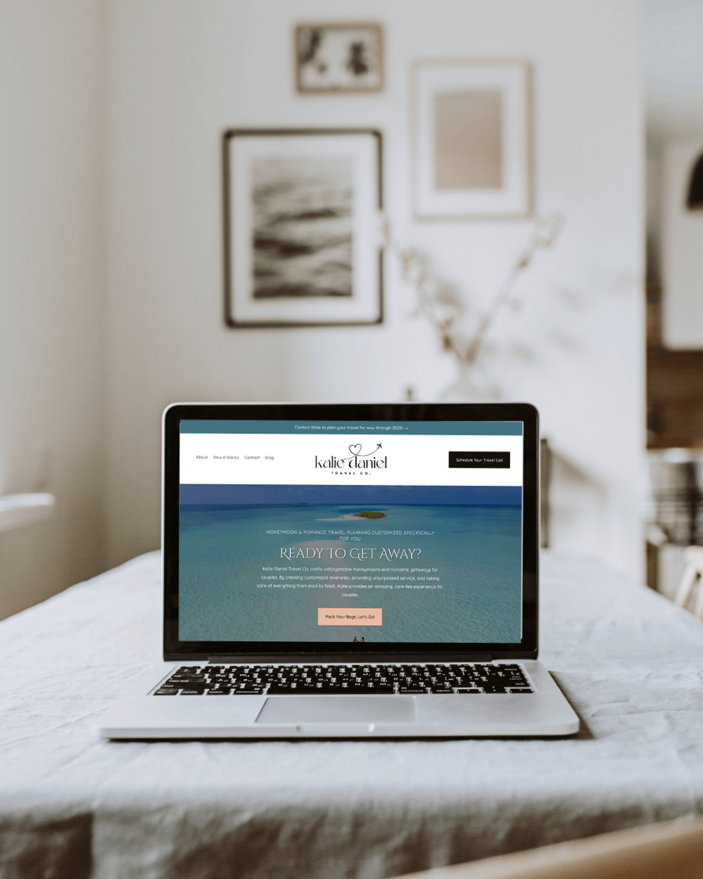A laptop on a table in a room with pictures hung on the wall behind it. The laptop features a web design by Las Vegas web designer Vivian Keithley of Desert Rose Design & Photo Co. for client Kalie Daniel Travel Co.